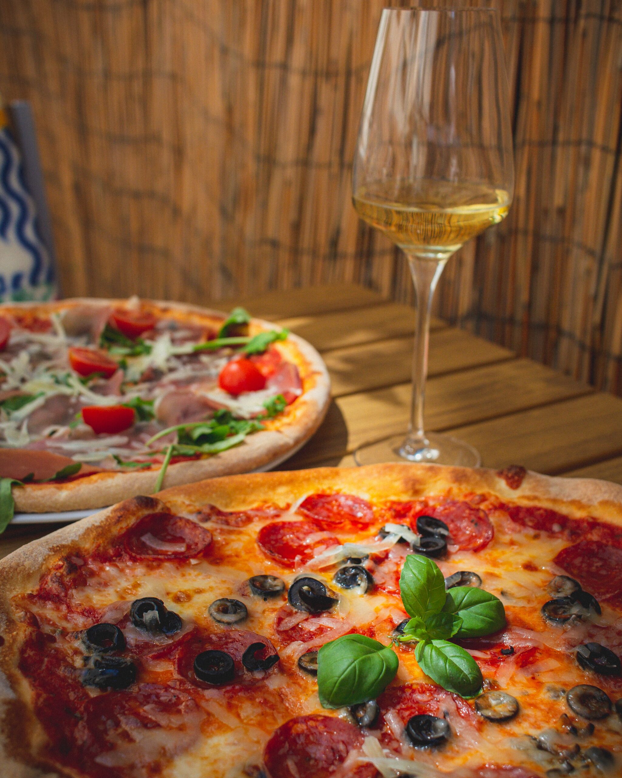 Close-up of gourmet pizzas with fresh toppings and a glass of white wine on a wooden table, perfect for food photography.